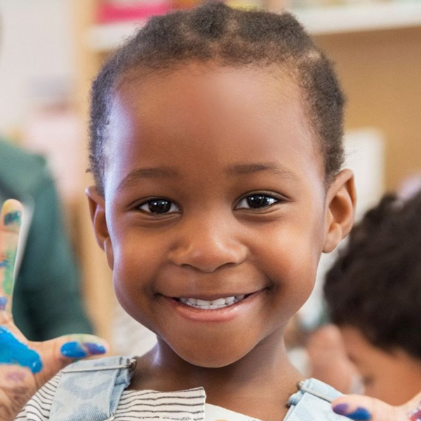 A young girl with colorful painted hands smiles in a vibrant classroom setting, showcasing her creativity and joy.